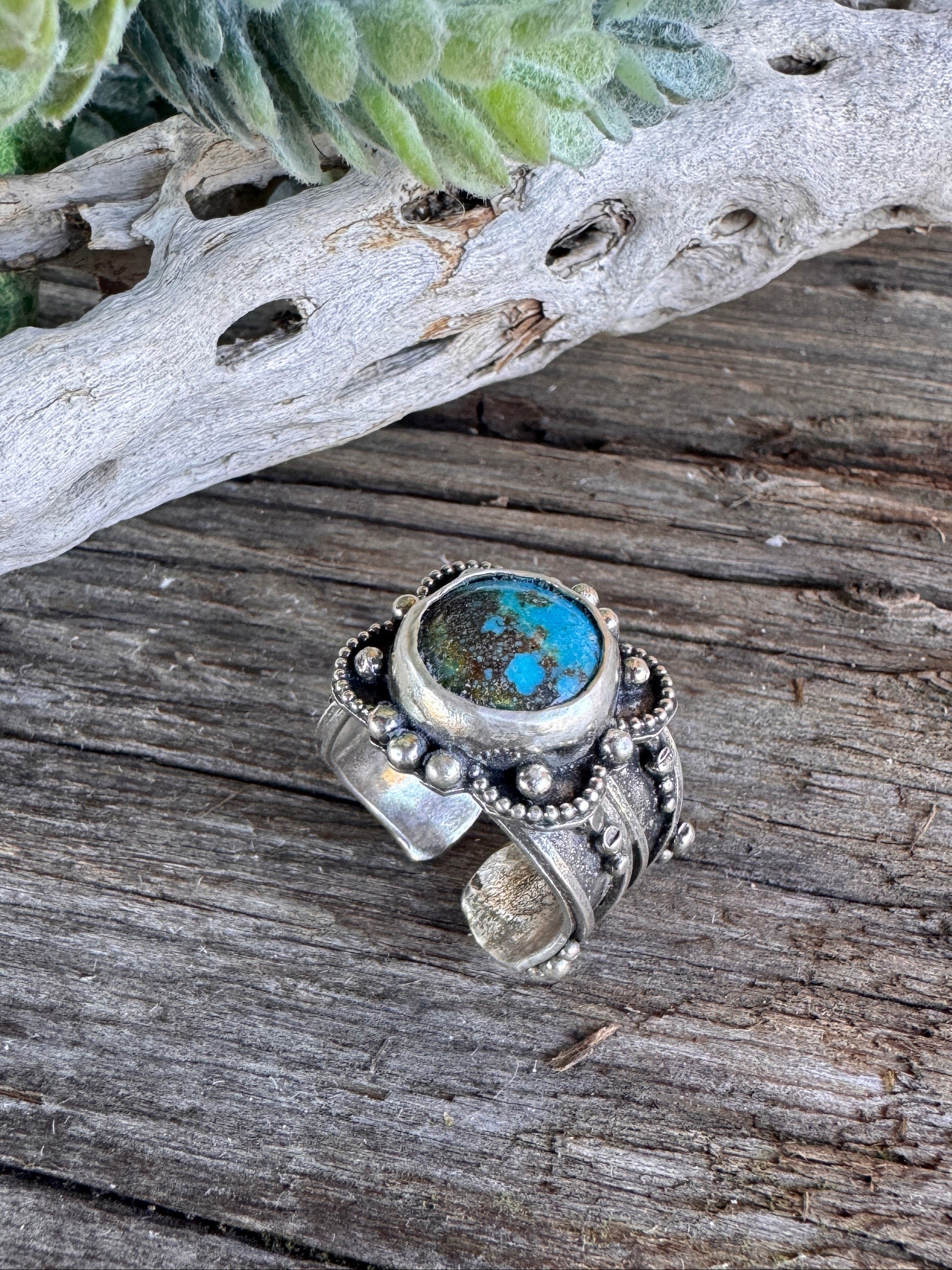 Desert Flower Turquoise & Silver ring with a central gemstone on a wooden surface with natural elements.