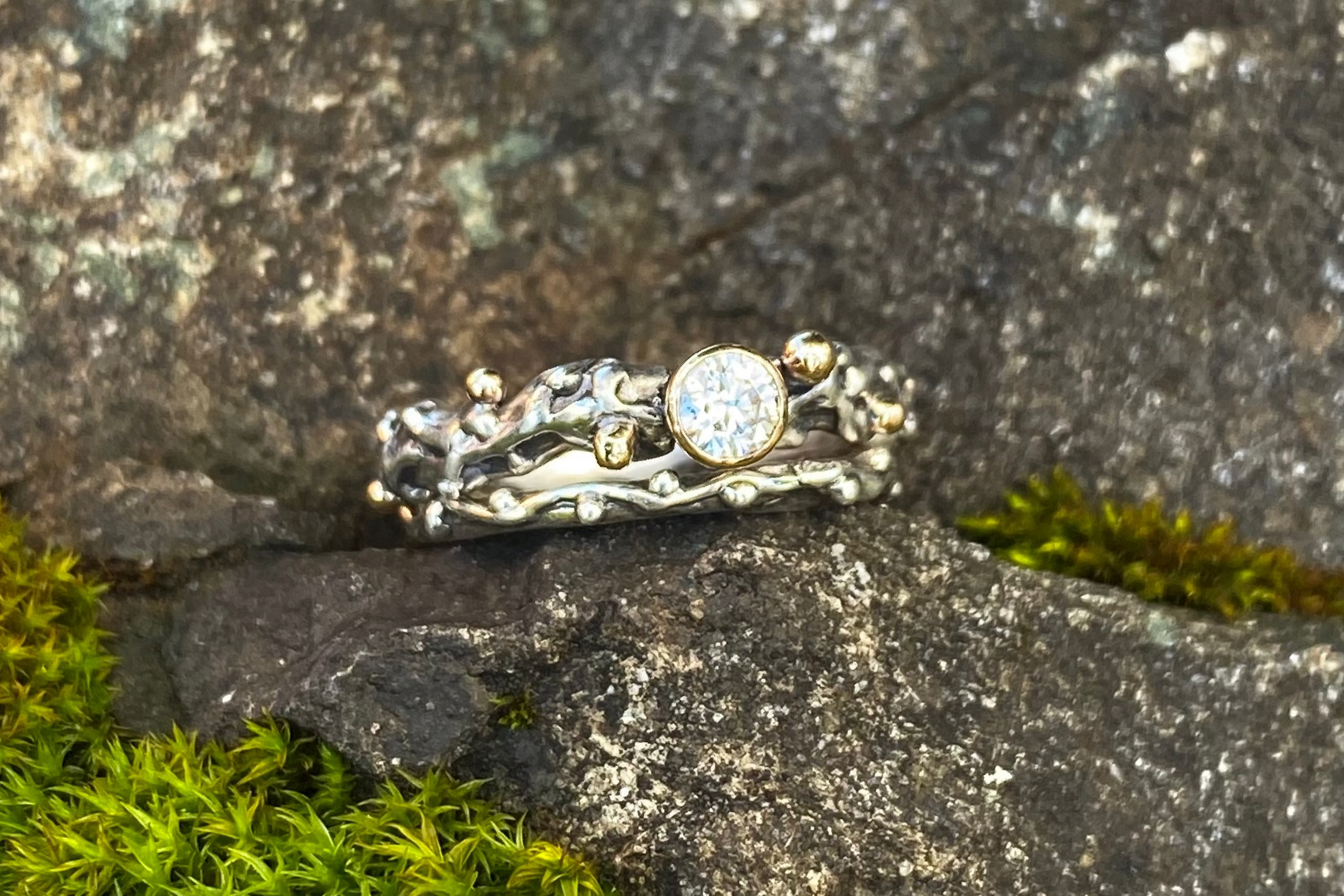 A double ring featuring a central moissanite gemstone set in a gold bezel, surrounded by silver vine-like pattern on a rocky surface with green moss.